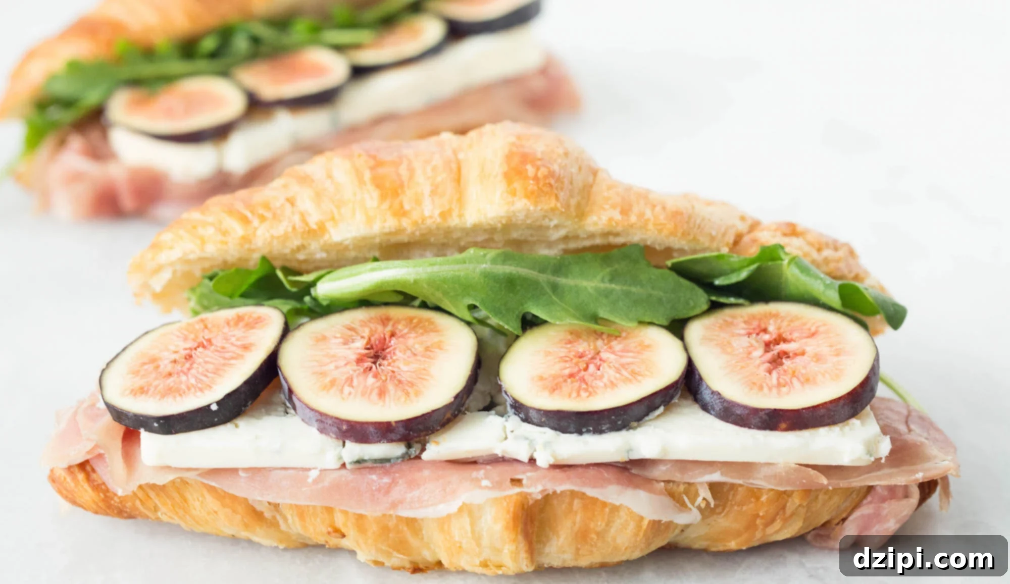 A croissant sandwich featuring prosciutto, blue cheese, sliced figs and arugula sits on a white background. Another fig sandwich is out of focus behind it.