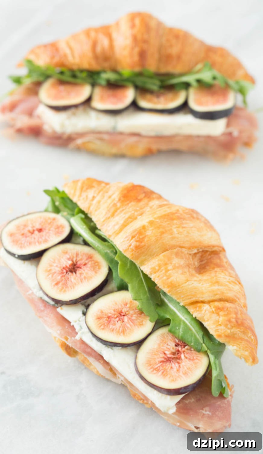 Two fig and blue cheese sandwiches sit on a white background. The croissant tops are positioned slightly off to the side so you can see the layers of prosciutto, blue cheese, sliced figs, and arugula. 