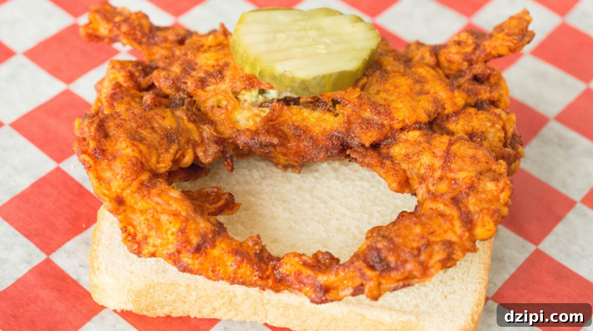 A soft shell crab that's been breaded, deep fried, and coated in Nashville hot sauce sits on a piece of white bread. There is a single pickle on top.