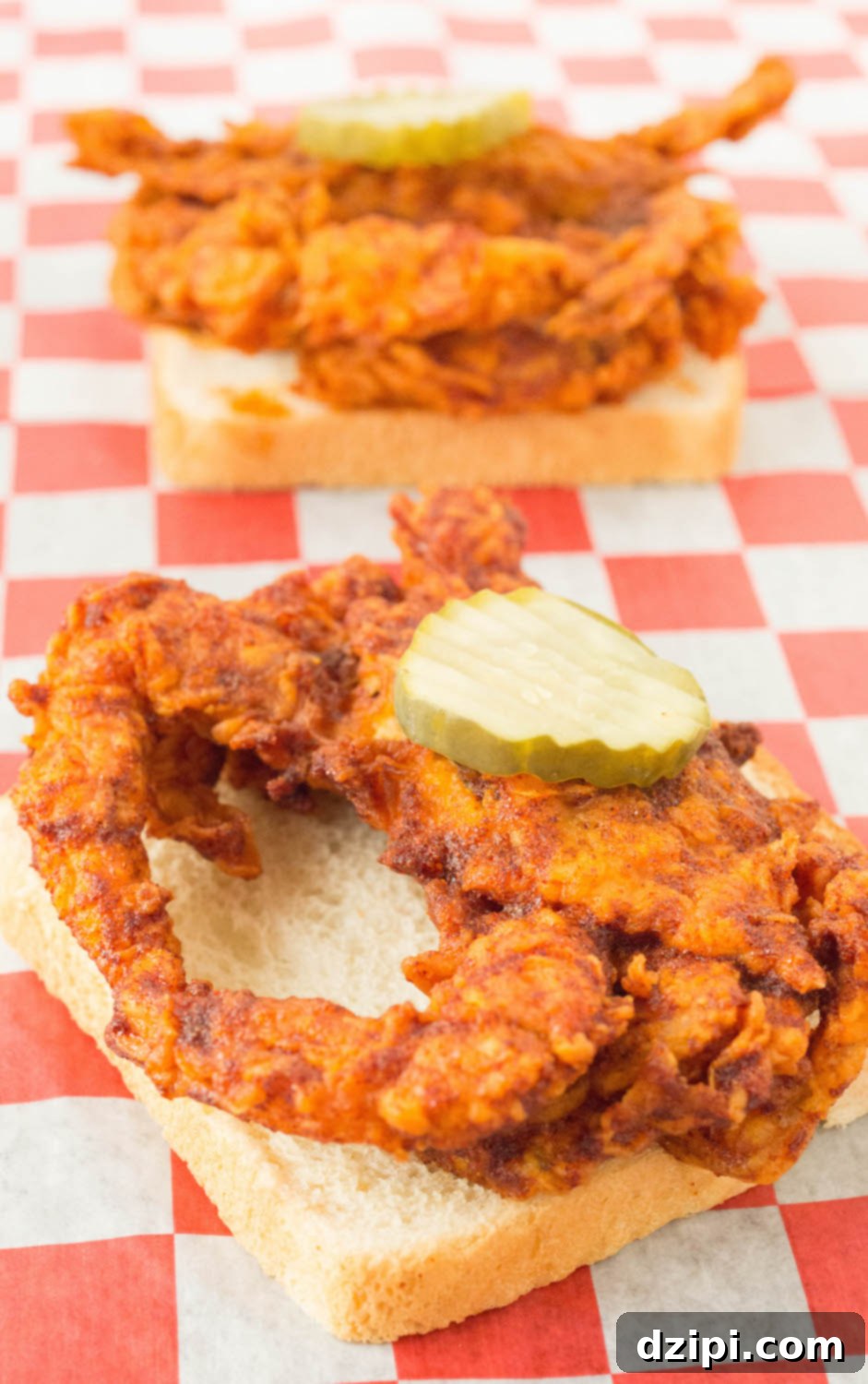 A deep fried soft shell crab that's been covered in Nashville hot sauce. It's sitting on top of a slice of white bread and has a pickle on top. Another one can be seen in the background.
