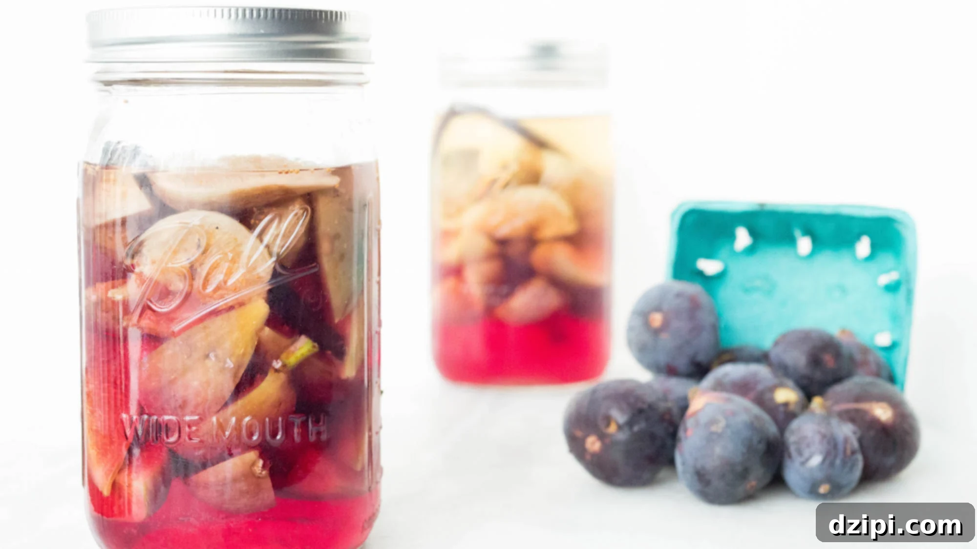 Homemade Fig Vodka Elixir 2 A large glass mason jar is filled with vodka that's turned a beautiful pink hue from the sliced figs inside. Another jar is out of focus in the background along with a basket of fresh brown figs, showcasing the delightful outcome of fig infusion.