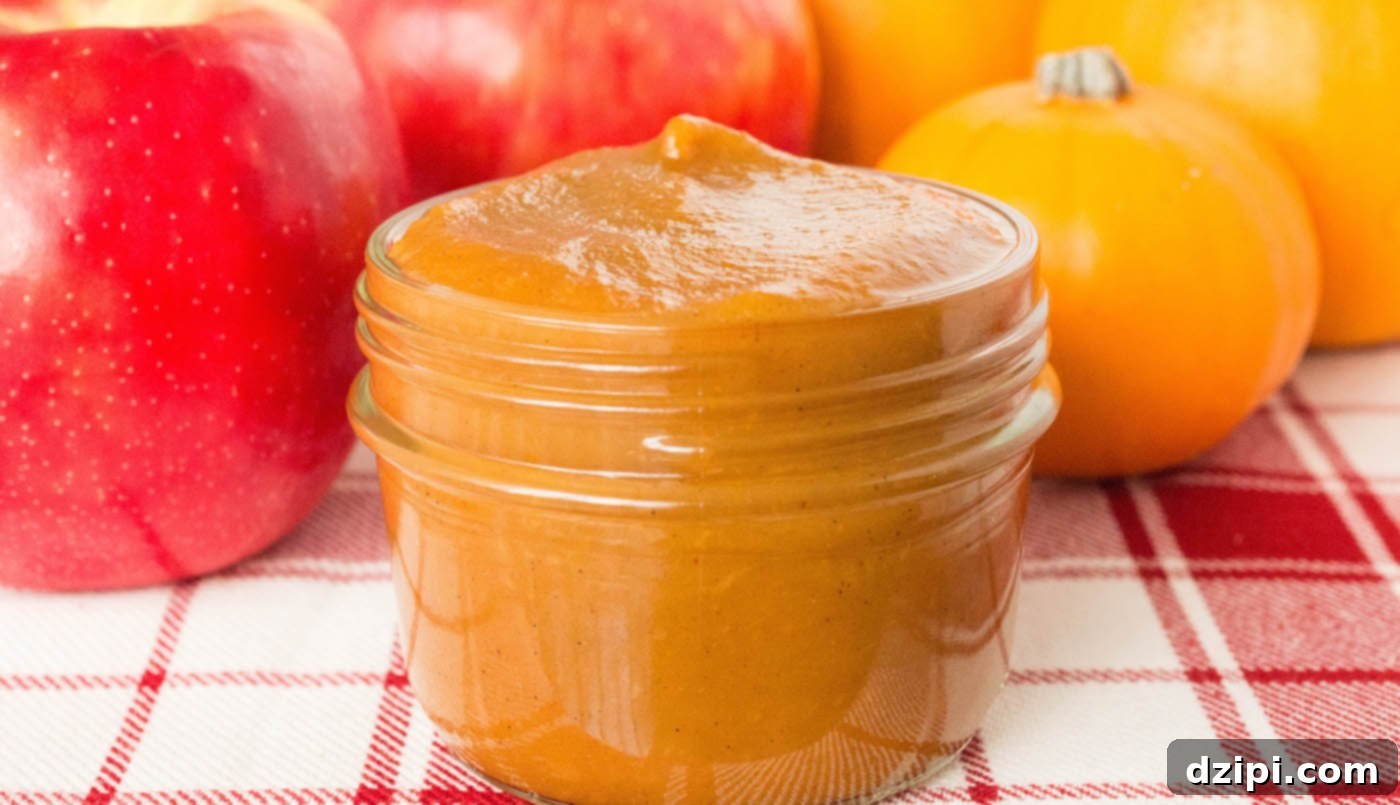 A small glass mason jar filled with rich, homemade pumpkin apple butter in focus. Out of focus in the background are small decorative pumpkins and a few fresh apples, setting a cozy autumn scene.