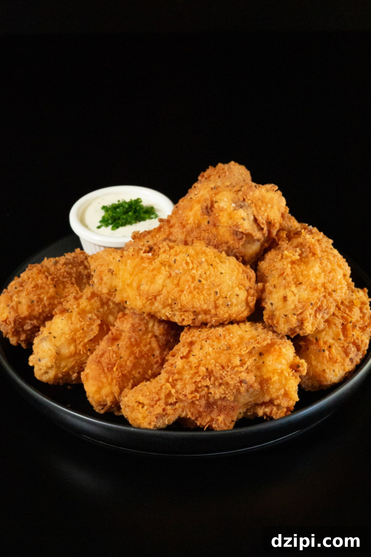 A small black plate holds a stack of about a dozen breaded and fried chicken wings. A small side of ranch sauce topped with chives is out of focus in the background, ready for dipping.