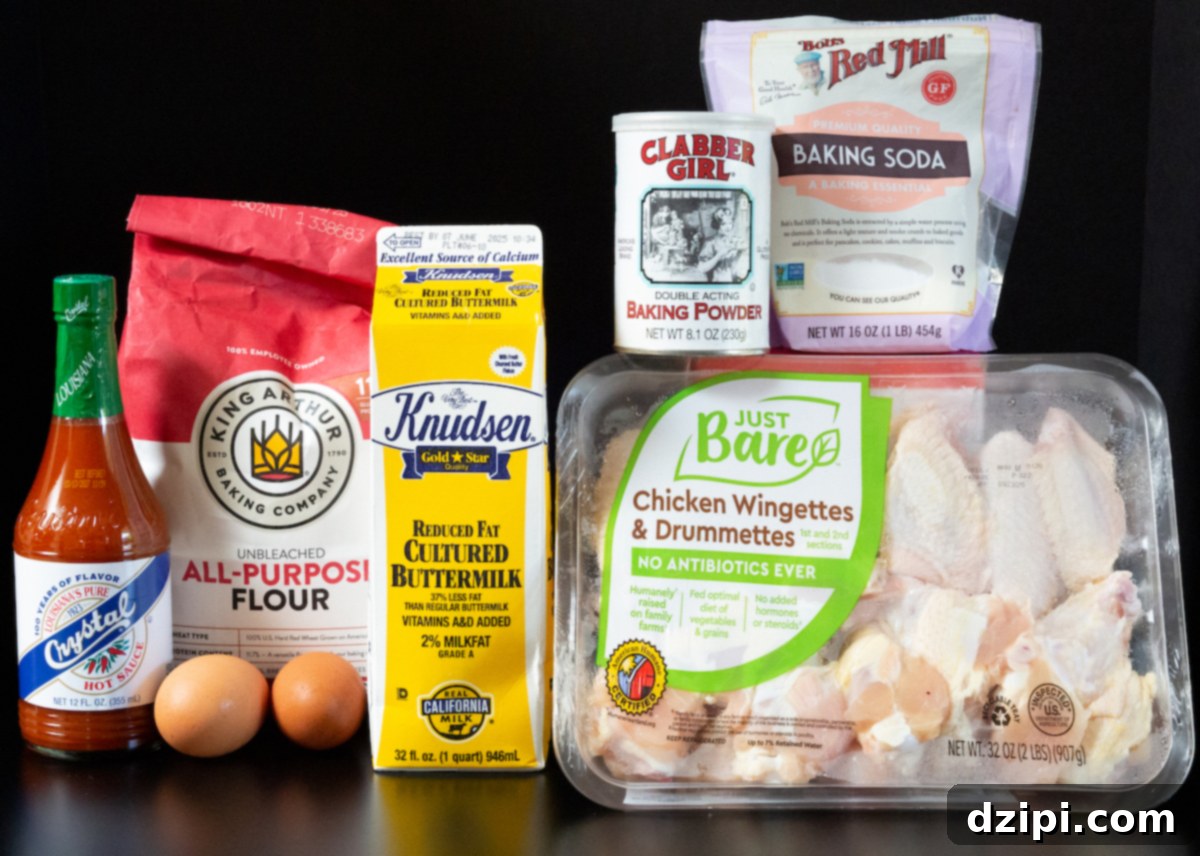 An overhead shot displays the essential ingredients for buttermilk fried chicken wings on a dark surface: raw chicken wings, a carton of buttermilk, all-purpose flour, baking soda, baking powder, two eggs, and a bottle of Crystal hot sauce.