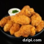 A small black plate has about a dozen breaded and fried chicken wings stacked on top of each other. A small side of ranch sauce topped with chives is out of focus in the background.