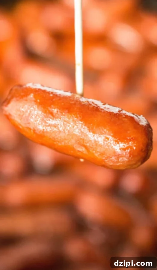 Spicy Apple Cranberry Glazed Little Smokies 4 Close up view of a toothpick holding a single apple cranberry little smokie, with a crock pot full of them blurred in the background.