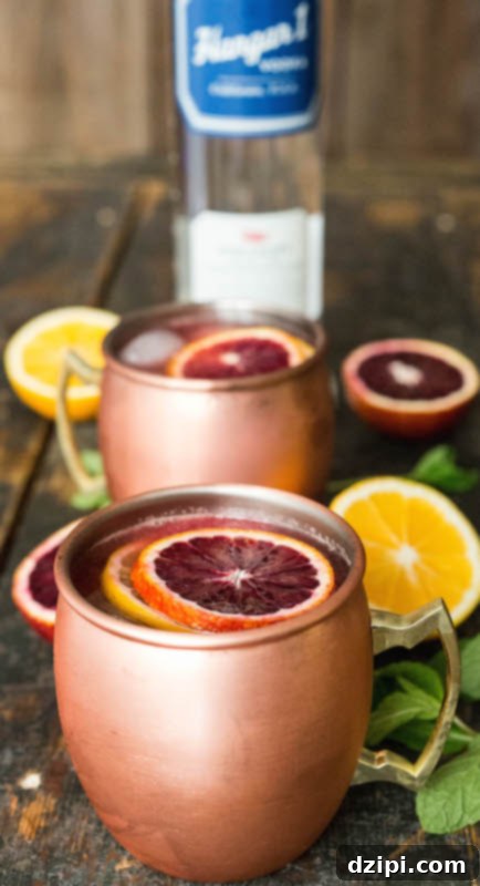 A glistening copper mug brimming with a blood orange & Meyer lemon Moscow Mule, elegantly garnished with a fresh slice of each citrus. Another mule and a premium vodka bottle subtly blur in the background, hinting at the ingredients.