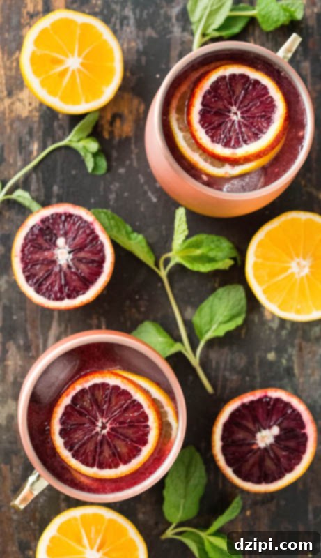 A stunning overhead shot featuring two copper mugs, each holding a beautifully crafted blood orange & Meyer lemon Moscow Mule, surrounded by fresh mint leaves and artfully arranged slices of blood orange and Meyer lemon.