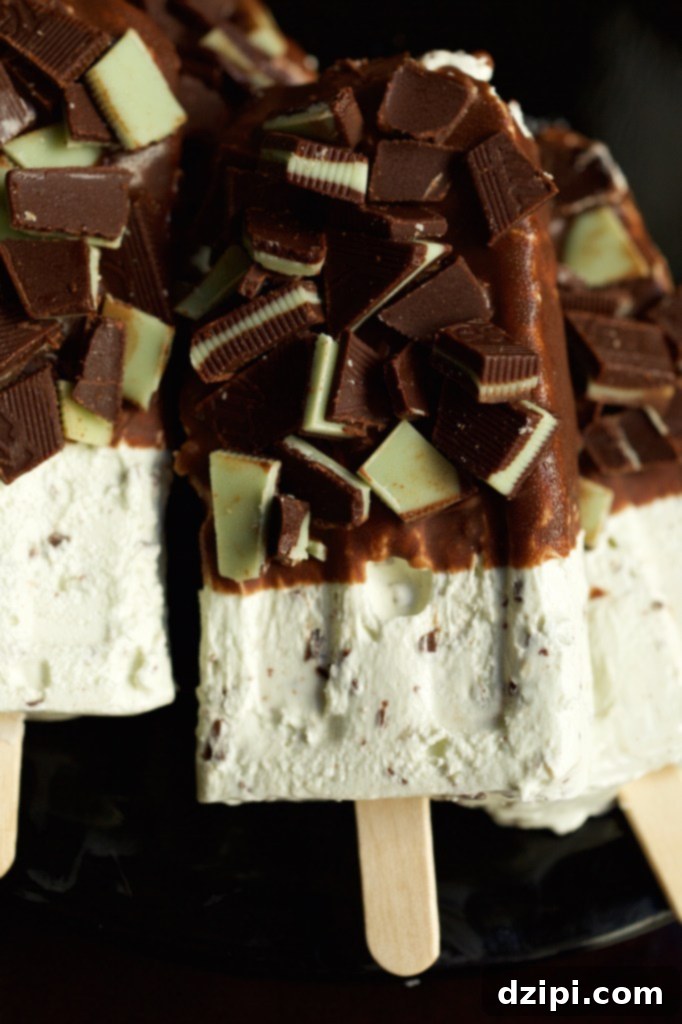 Cool Mint Chocolate Cheesecake Popsicles 4 A close-up image of a single mint chocolate cheesecake popsicle. The bottom half reveals its light green, creamy cheesecake interior, while the top is generously coated in a dark chocolate shell and adorned with sparkling green and brown chopped Andes mints.