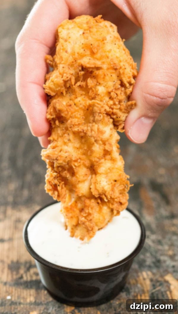 A freshly fried buttermilk chicken tender being generously dipped into a creamy ranch dressing.