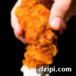 A close-up of a hand holding a homemade buffalo chicken tender that's been dipped in ranch sauce against a black background